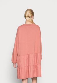 Vaaleanpunainen pitkähihaisessa mekossa on kerroksellinen röyhelöhelma. Pehmeä kangas, rento malli, pyöreä pääntie ja tilavat hihat. Takakuvassa.