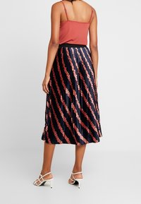 Kvinna som står klädd i en ärmlös topp i rostfärgad nyans, en plisserad marinblå kjol med rosa diagonala ränder och vita högklackade sandaler.