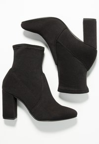 Botas negras de tobillo hechas de tela suave, con punta afilada y tacón ancho, que presentan un diseño sin costuras y mínimos acentos de metal.