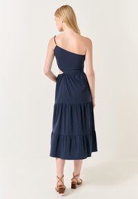 Navyblaue Baumwoll-Midikleid mit einem schulterfreien Träger, gestuftem Volantsaum, Ausschnitt und verstellbarem beaded Träger. Braune Sandalen mit Absatz.