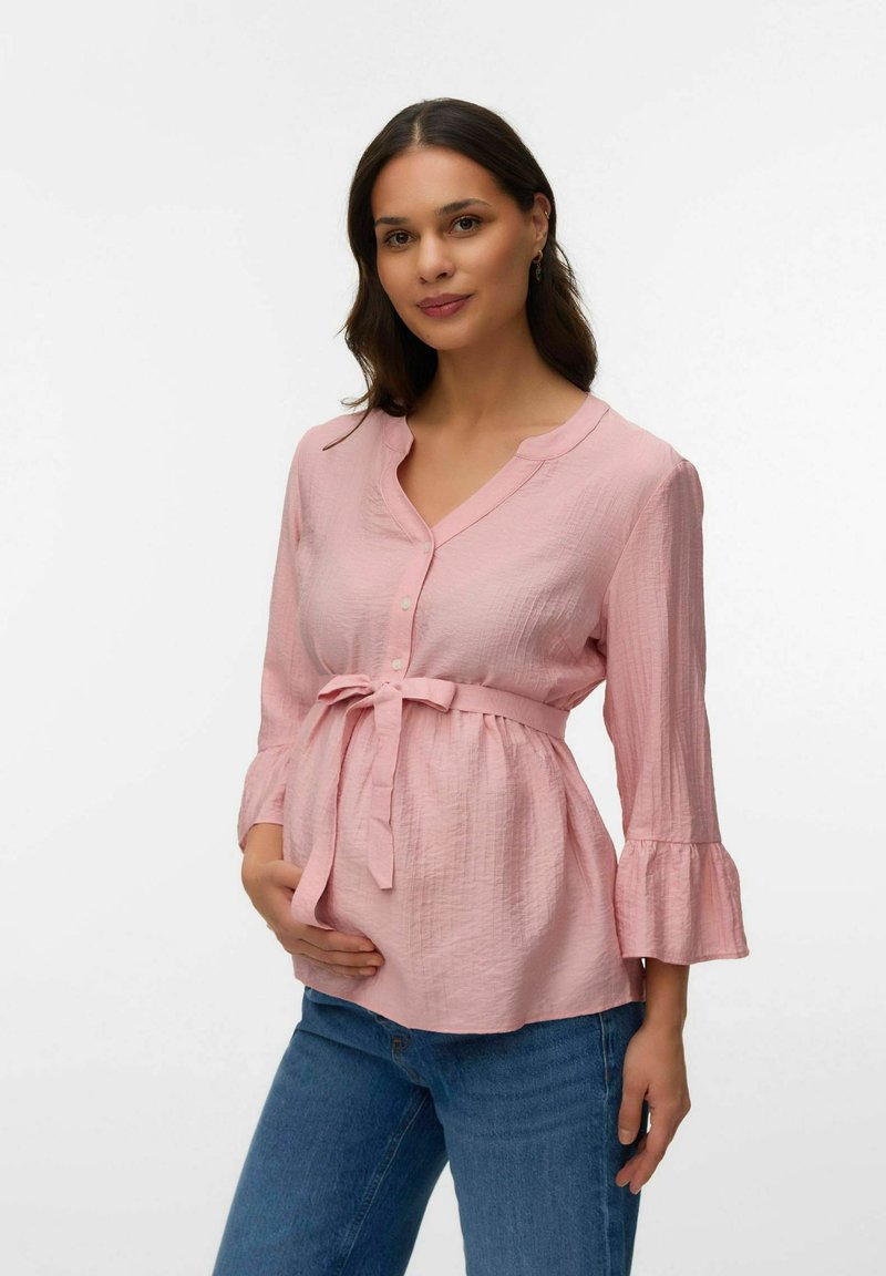 Rase ema roosas nööbitud pluusis, millel on vöökohal pael, hoides kõhtu, siniste teksadega, seisab tavalise valge tausta ees.