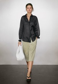 Camicia nera in lino con bottoni, maniche lunghe leggere, abbinata a pantaloni corti verde chiaro e sandali neri. Il modello tiene una borsa bianca intrecciata.