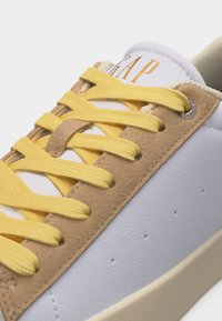 Gros plan d'une chaussure de sport blanche et beige avec des lacets jaunes et un logo doré "GAP" sur la languette, montrant en détail les coutures et les œillets.
