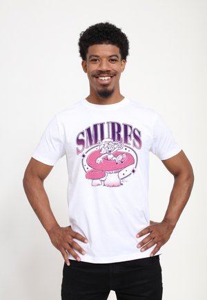 Man smiling wearing a white t-shirt with a pink Smurf sitting on a large mushroom and "SMURFS" text above.