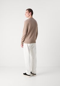 Maglione beige a maglia con scollatura rotonda, indossato con pantaloni bianchi e scarpe slip-on nere, visto da dietro su uno sfondo semplice.