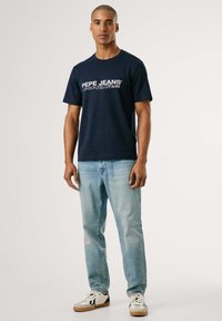 Marineblauw katoenen T-shirt met witte "Pepe Jeans" tekst, gecombineerd met lichtblauwe spijkerbroek en witte sneakers met rode en marineblauwe accenten.