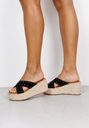 Piernas con sandalias cuña negras con tiras cruzadas y tachuelas, suela de plataforma beige texturizada sobre suelo gris.