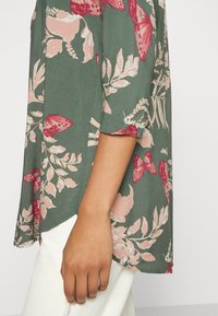 Bloemige blouse van donkergroene stof, versierd met roze bloemen en vlinders, met korte mouwen en een losse pasvorm. Randen zijn zacht afgewerkt.