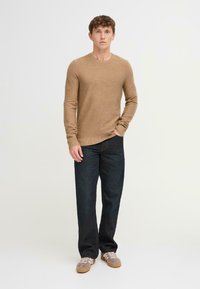 Suéter de punto marrón claro con cuello redondo, combinado con jeans de mezclilla oscuros. El modelo también lleva zapatillas grises con cordones blancos.
