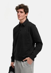 Schwarzer Pullover mit Kragen, ausgestattet mit einer Vorderblende, zwei Knöpfen und einem kleinen Logo auf der linken Brust. Der Stoff scheint weich zu sein.