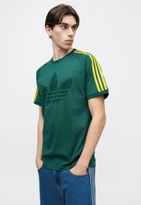 Jauns vīrietis melnā Adidas T-kreklā ar dzeltenām svītrām uz piedurknēm un lielu trešo logotipu, rokās kabatās, skatās uz sāniem.