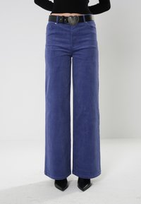Pantalones de pierna ancha morados, hechos de material suave, con un cinturón negro con hebilla en forma de corazón, combinados con zapatos de punta negros.