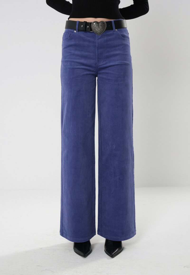 Pantalones de pierna ancha morados, hechos de material suave, con un cinturón negro con hebilla en forma de corazón, combinados con zapatos de punta negros.