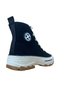 Zapatilla alta negra con cordones blancos, suela blanca gruesa, piso marrón y parche redondo en el tobillo que dice "The Urban Land."