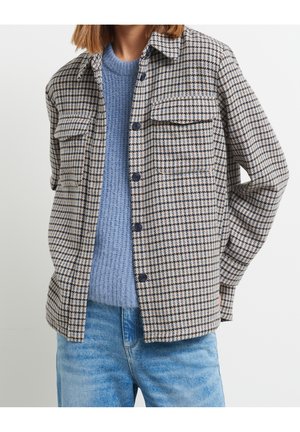 Persona con suéter de punto azul, vaqueros azules y una chaqueta abierta de cuadros con bolsillos en el pecho