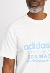Valkoinen puuvillainen t-paita, jossa on vaaleansinistä painettua tekstiä ja pieni beige Adidas-logo vasemmassa rinnassa. Pyöreä pääntie ja lyhyet hihat.