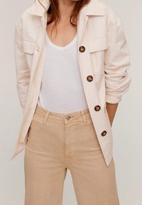 Giacca color crema con colletto, tasche frontali e bottoni marroni, indossata sopra una maglietta bianca e pantaloni beige. Texture del tessuto liscia.