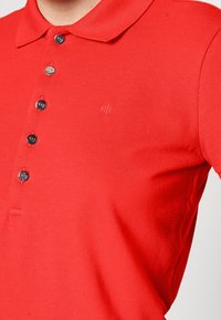 Röd polo-shirt med fem knappar, liten broderad "RL" logo på bröstet och en klassisk krage synlig på en person.