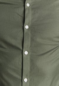 Vihreä nappipaita, joka on valmistettu teksturoidusta kankaasta. Paidassa on suora leikkaus ja edessä valkoiset napit.