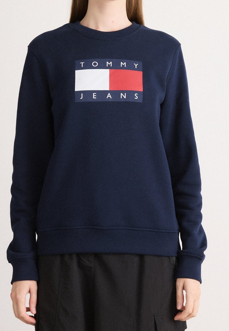 Sweat bleu marine avec un logo rectangulaire blanc et rouge et le texte "TOMMY JEANS" sur la poitrine, porté par une personne aux cheveux longs.