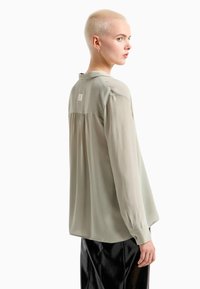 Camicia verde chiaro a maniche lunghe, realizzata in tessuto liscio. Presenta un colletto arrotondato, un dettaglio arriccicato sul retro e un piccolo logo nella parte superiore della schiena.