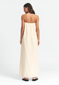 Trägerloses, cremefarbenes Maxikleid aus strukturiertem Material. Verfügt über ein gerafftes Oberteil und reicht bis zu den Knöcheln, kombiniert mit schwarzen Sandalen.