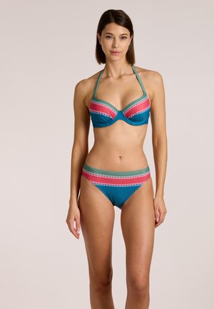 Frau trägt einen türkisfarbenen Bikini mit rosa, grünen und weißen horizontalen Streifen am Rand, steht vor einem schlichten beigen Hintergrund.