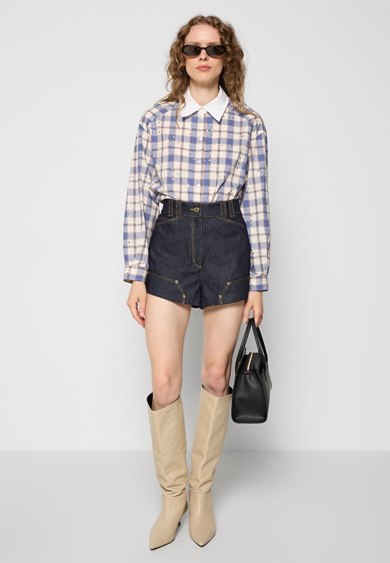 Camisa de cuadros de manga larga con un collar blanco, combinada con pantalones cortos de mezclilla de cintura alta. La modelo lleva botas beiges hasta la rodilla y sostiene un bolso negro.