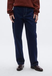 Pantalones de mezclilla azul oscuro con costuras blancas contrastantes, corte recto, bolsillos laterales y un ajuste relajado. Combinados con zapatos marrón oscuro.