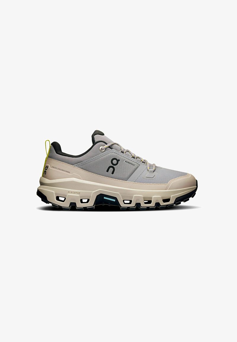 Hellgraue Sportschuh mit einem Obermaterial aus Mesh, schwarzen Akzenten, gepolstertem Kragen und einer markanten dicken Sohle mit Belüftungsöffnungen.
