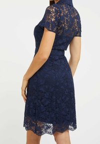 Robe en dentelle bleu marine à manches courtes, avec des motifs floraux et une silhouette ajustée. Détail décoratif à l'ourlet festonné de la jupe.