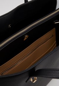 Intérieur de sac à main texturé noir avec une poche zippée et des compartiments ouverts, doté d'une fermeture éclair et de boutons en métal doré.