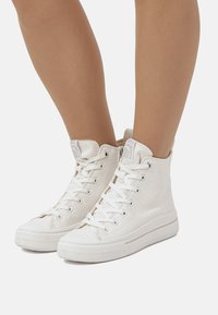 Sneaker alte bianche in tela con lacci bianchi, suole in gomma e un'etichetta con logo sulla parte superiore. Presentano una superficie in tessuto testurizzato e un design classico.