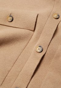 Tessuto di lana beige con una texture liscia, dotato di un colletto e tre bottoni rotondi a quattro fori nella parte anteriore. Dettagli di cucitura puliti e visibili.