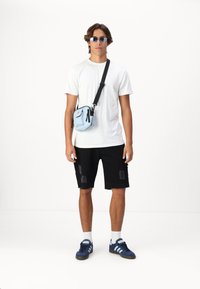 Bolso cruzado azul claro con cremallera, llevado sobre una camiseta blanca, pantalones cortos rasgados negros y zapatillas deportivas azules con tres rayas azul claro.