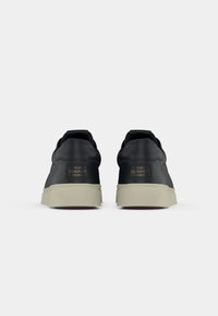 Sneakers din piele neagră cu talpă din cauciuc crem, dotate cu un accent logo auriu pe călcâi și design minimalist. Textură netedă.