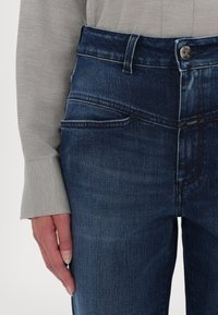 Jean en denim bleu foncé avec poches avant et fermeture par bouton, porté avec un haut à manches longues gris clair, main reposant le long du corps.
