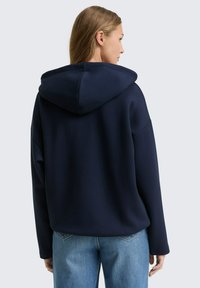 Donna di spalle che indossa un hoodie blu navy e jeans azzurri su uno sfondo grigio chiaro uniforme.