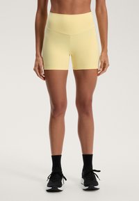 Rumene atletske hlače z visokim pasom, prilegajoča zasnova, gladka tekstura in kontrastni črni atletski čevlji.