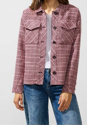 Frau trägt eine rot-weiße strukturierte Jacke mit Knöpfen über einem weißen Oberteil, kombiniert mit blauen Jeans, gezeigt von Hals bis Hüfte.