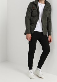 Oliven grøn, lynlåset jakke med flere lommer, båret over en hvid t-shirt, parret med sorte joggers og hvide sneakers.