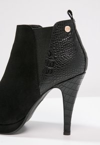 Botín negro con tacón stiletto alto. Presenta material de ante en la parte delantera y piel de cocodrilo sintética texturizada en la parte trasera.