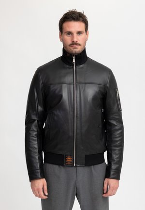 Man met kort donker haar en snor, die een zwarte leren jas met rits draagt, staand tegen een effen lichte achtergrond.