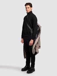 Čierna mikina so zipsom, zúžené nohavice a robustné čierne topánky. Na jednom ramene visí prešívaná bunda v neutrálnej farbe.