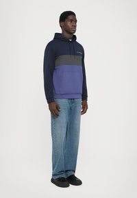 Um hoodie azul-marinho e roxo com um bolso frontal, risca cinza horizontal, capuz com cordão, combinado com calças de ganga de perna reta azul-claro e ténis pretos.