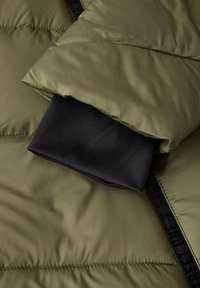 Olivgrön puffjacka i tyg med glans, med en svart inre manschett och en synlig logostropp längs dragkedjan. Mjuk och texturerad.