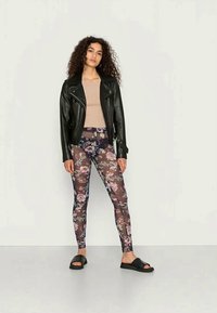 Jaca de motociclista em couro preto, blusa bege, leggings com padrão floral em tons escuros e sandálias de slide pretas. Cabelos cacheados, pose em pé.