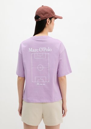 Γυναίκα που φορά ένα λιλά T-shirt με γραφικό λευκού γηπέδου ποδοσφαίρου και κείμενο "Marc O'Polo" στην πλάτη, μπεζ σορτς και κόκκινο καπέλο.