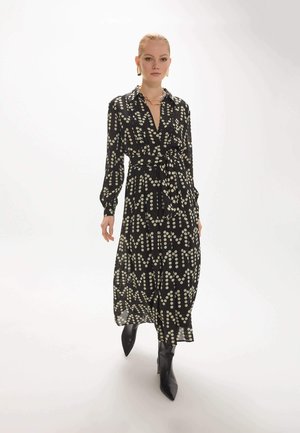 Femme portant une robe midi noire à manches longues avec des motifs légers en points, assortie à des bottines noires à talons, marchant sur un fond blanc uni.