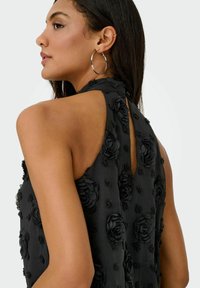 Femme portant une robe noire sans manches avec des motifs floraux en relief, des boucles d'oreilles en forme d'anneau et un bijou d'oreille, vue de dos et de profil.